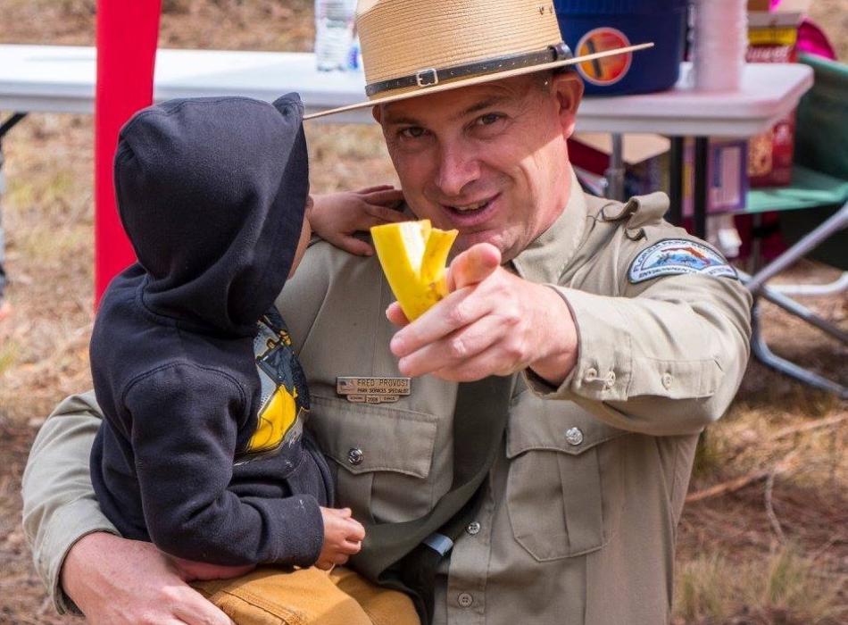 Park Services Specialist Fred Provost points at the camera while holding small child who is admiring his uniform. 