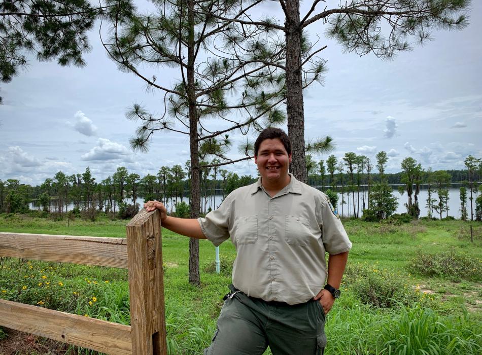 Park Ranger Sam Louzan smiling for the camera with pine trees and a lake behind him