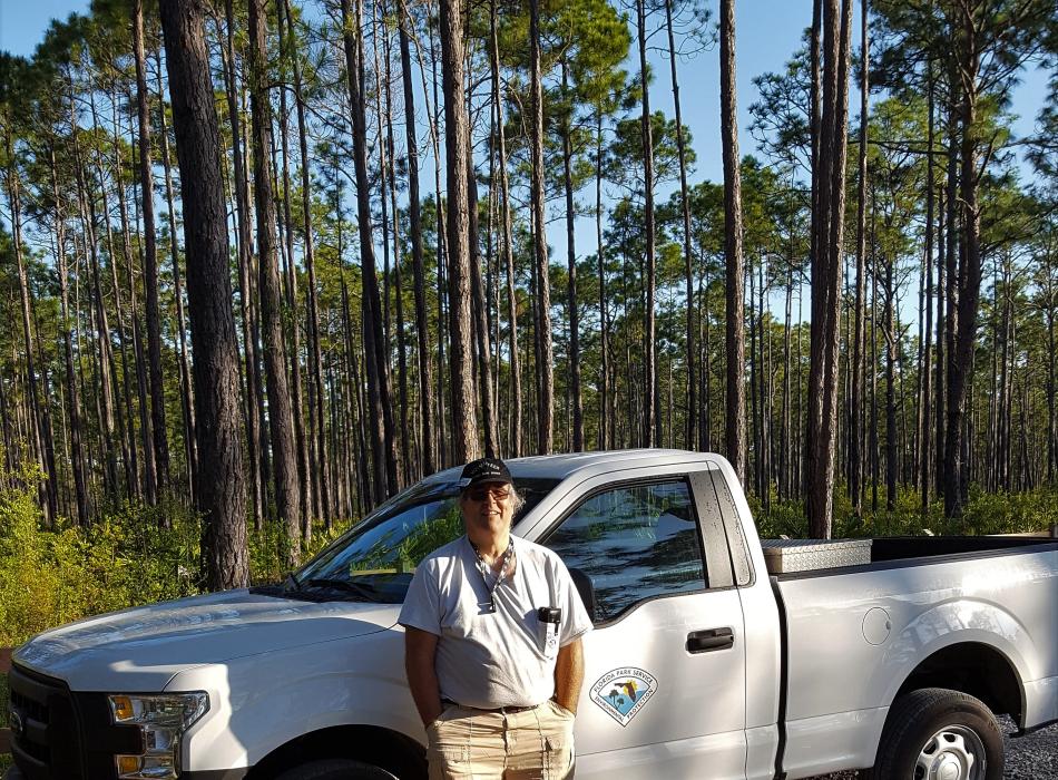 Volunteer Jeff Junior stands in front of park truck and forest of trees. 