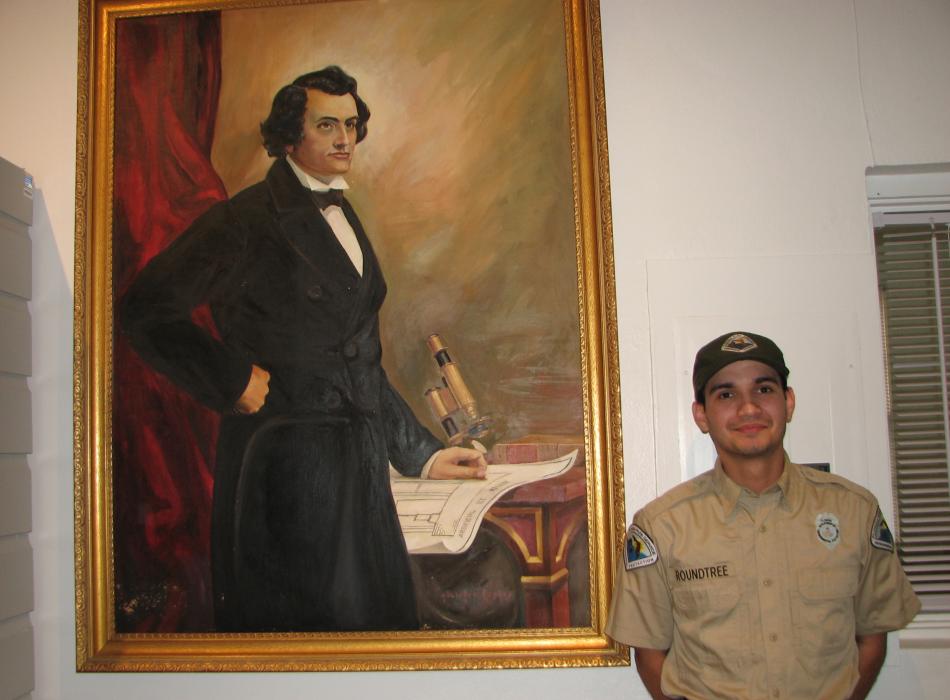 Park Ranger Jeromy Roundtree stands next to large painted portrait of John Gorrie displayed in the museum.