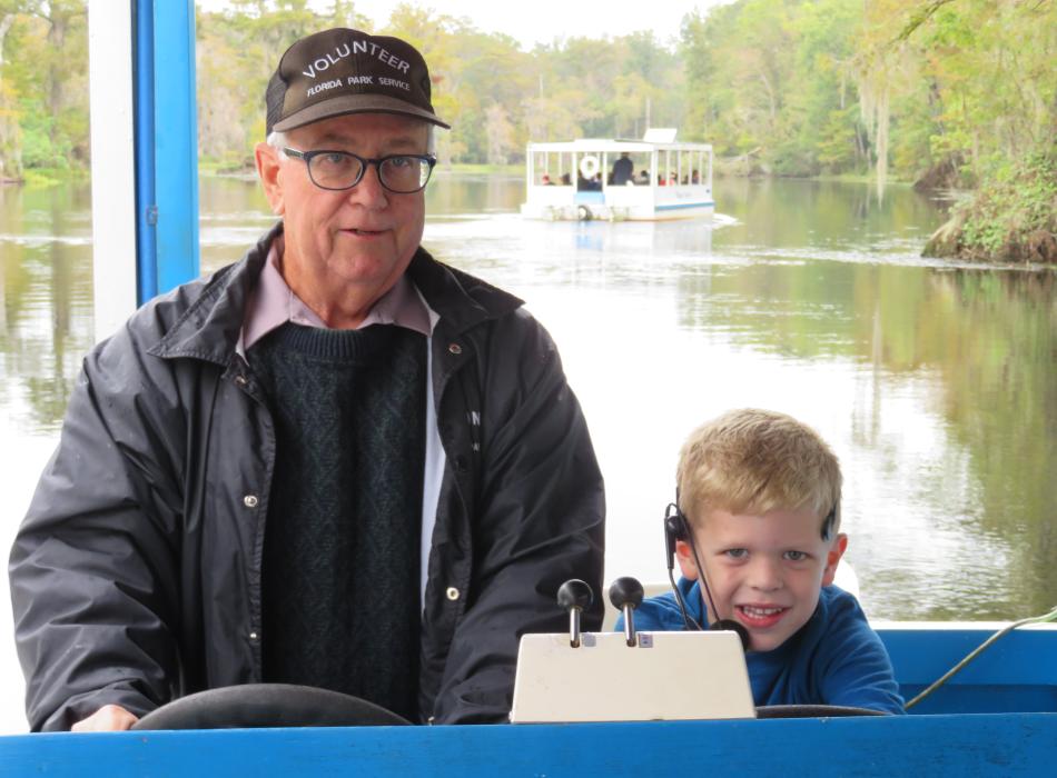 Volunteer at captains wheel of tour boat with young boy sitting nearby as co-captain. River boats can be seen on the river in the distance.