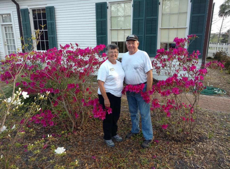 Volunteers Ken and Pattie Fox stand in front of azalea bushes at the Orman House full of bright fuschia blooms. 