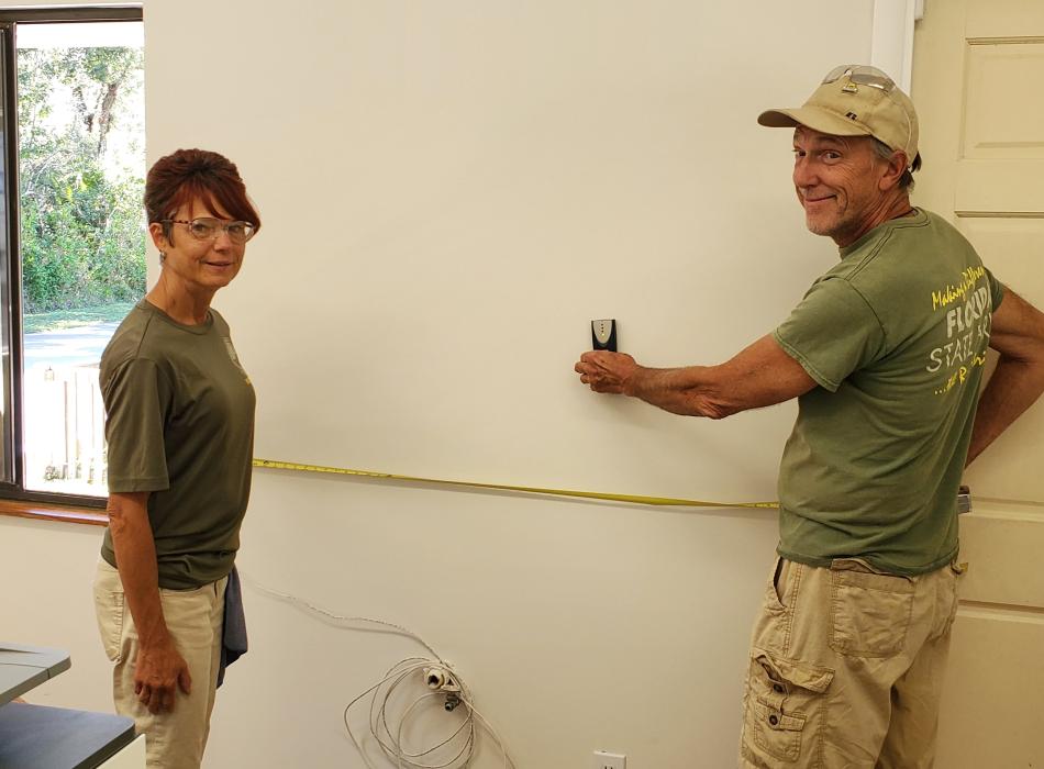 Two people measuring wall wearing green shirts and tan pants