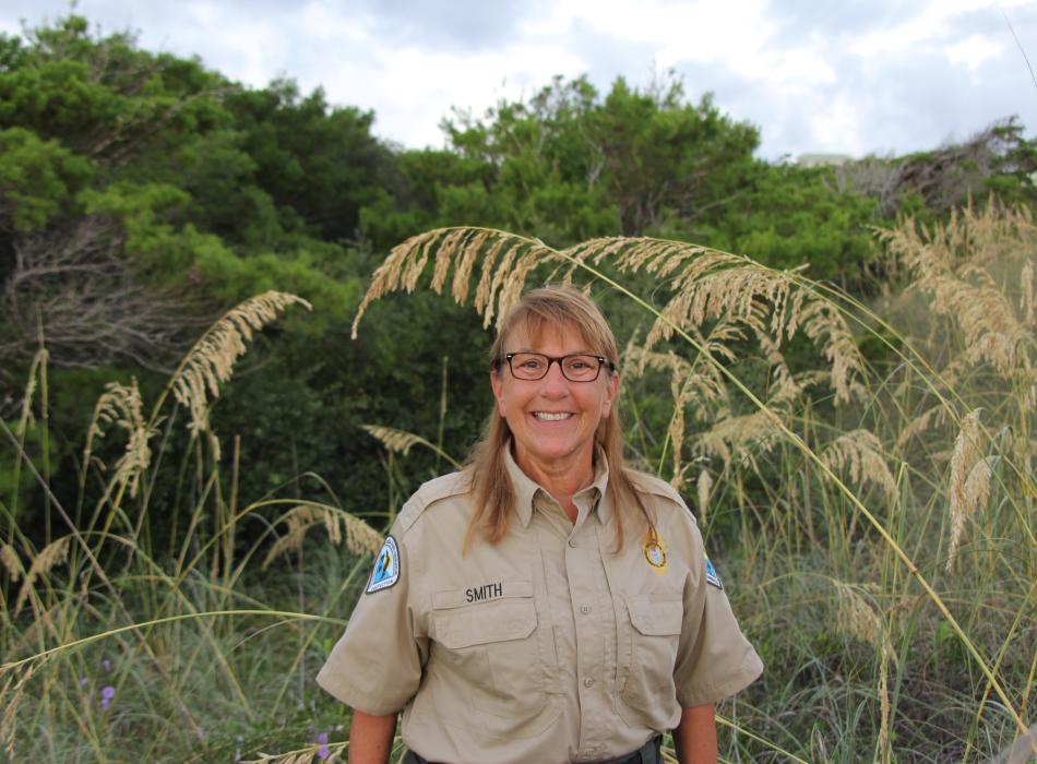 Lynda, smiling at the camera, standing in field of greenery.