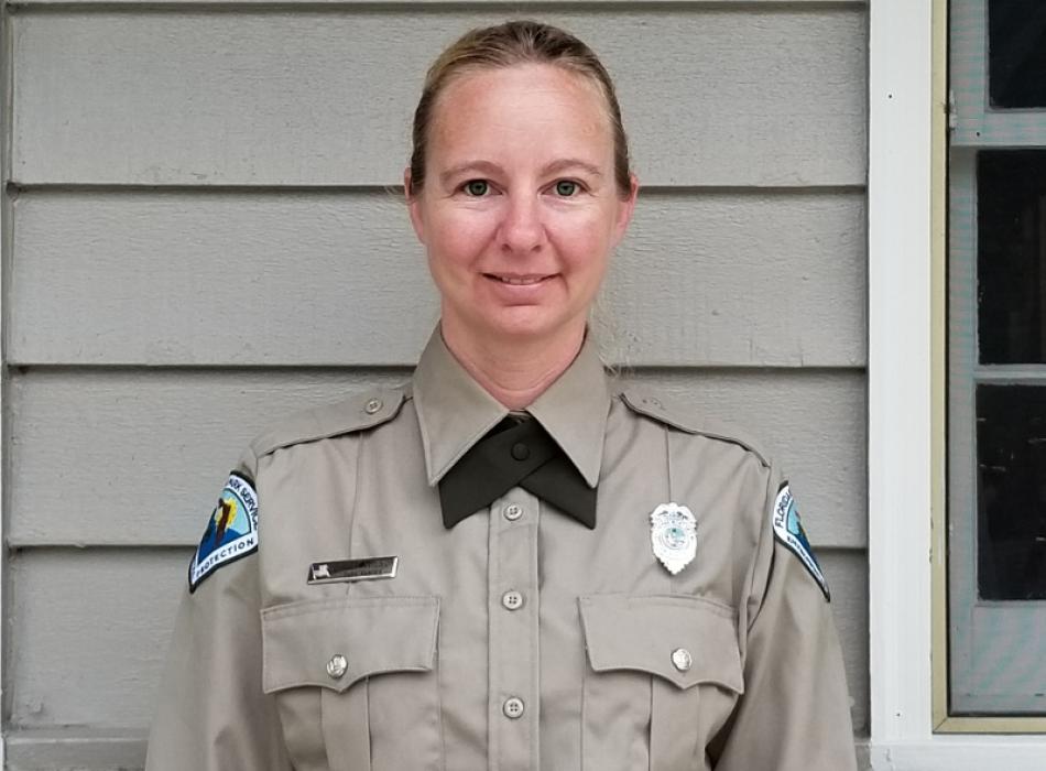 a woman stands against a park building in full uniform, smiling at the camera.