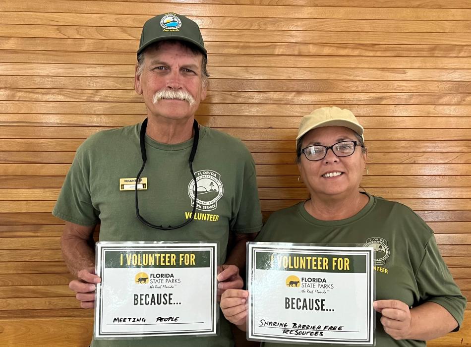 Pam and James hold up signs about why they volunteer: "meeting people" and "sharing barrier free resources."