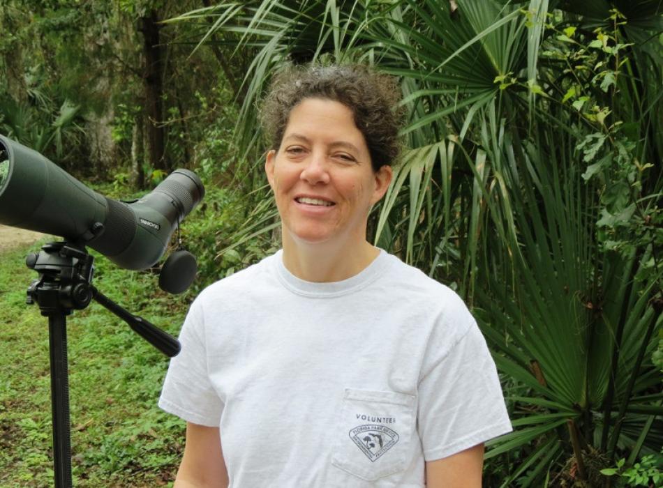 a woman in a volunteer shirt stands next to a spotting scope smiling
