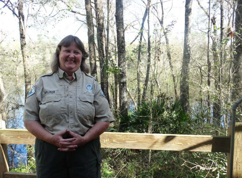 a woman in a park service uniform stands smiling in front of a wet forest.