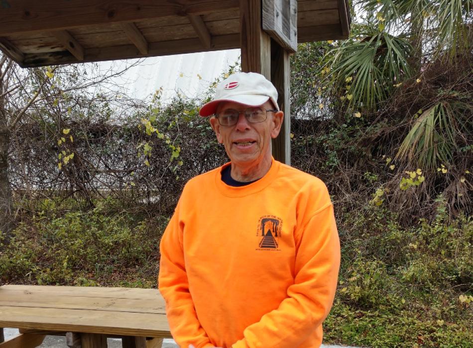 A man in an orange shirt and glasses smiles beside a bench.