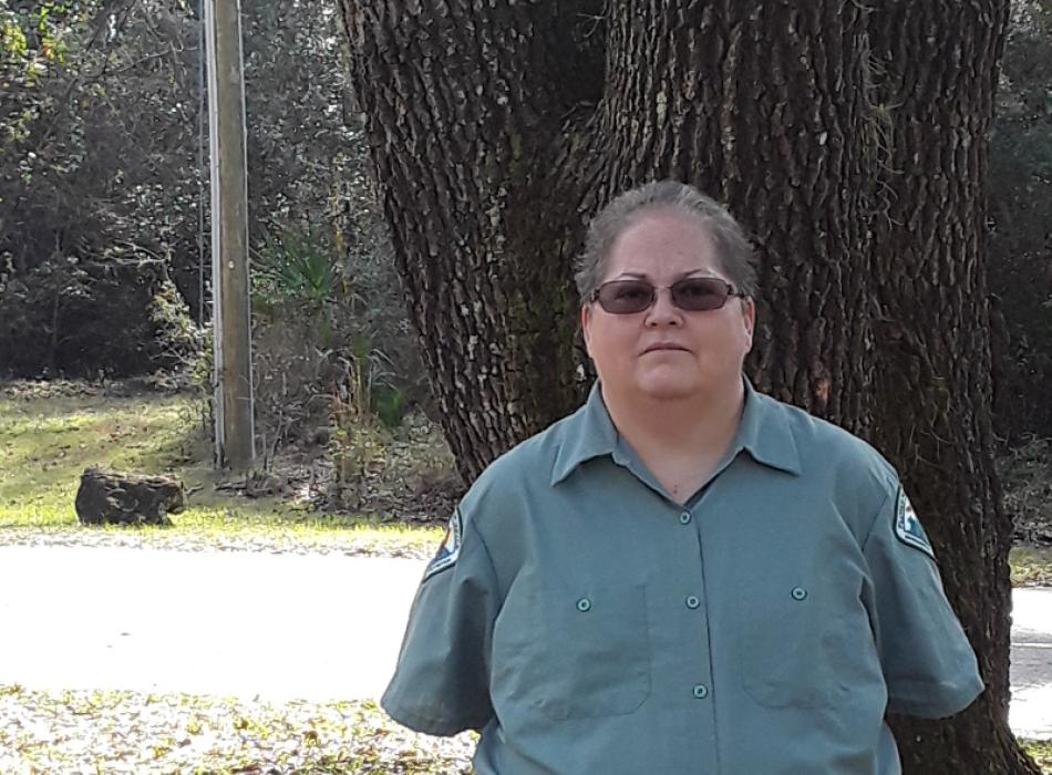 a woman with short hair and in a green uniform stand sin front of a tree