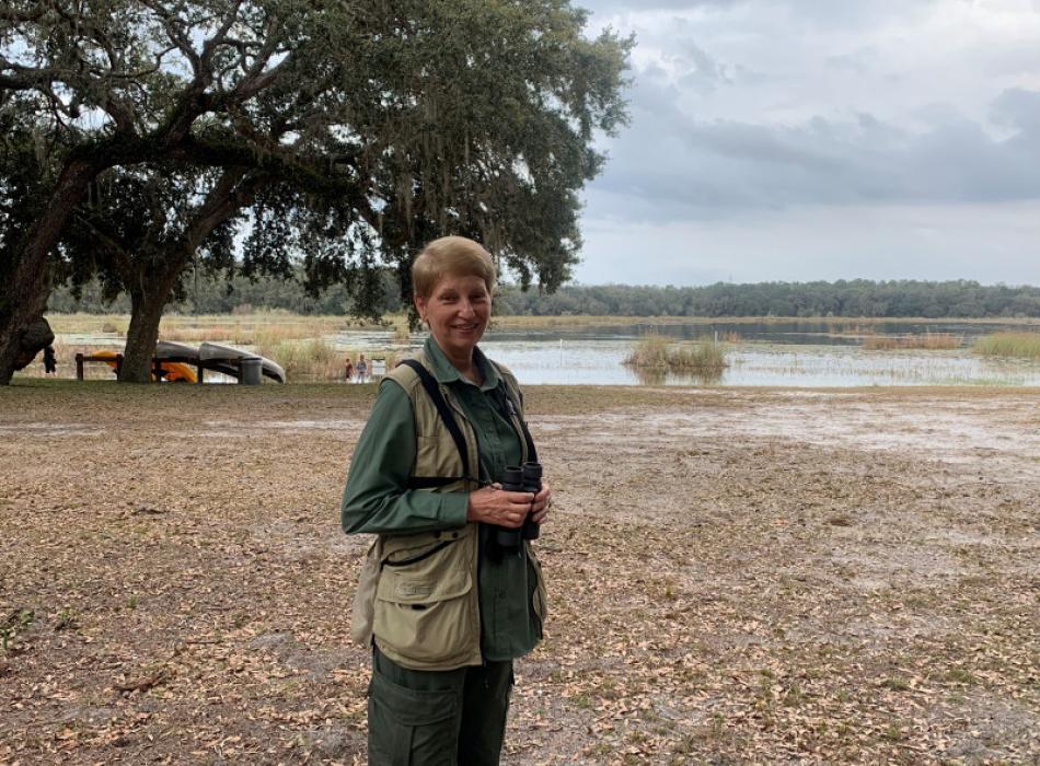 a woman stands beside a lake with binoculars.