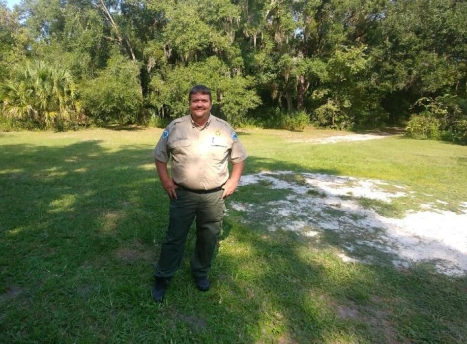 A man in a uniform stands in front of trees.