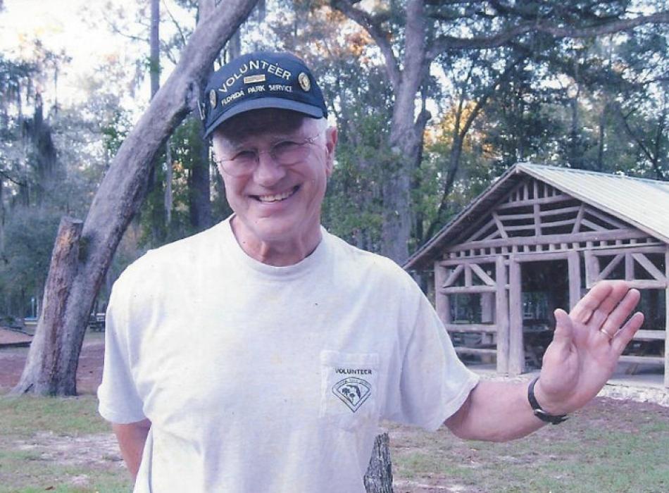 a man in a volunteer shirt and hat smiles and waves in front of a log pavilion