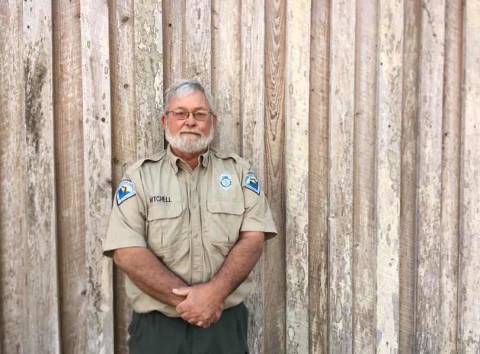 a man in a park service uniform stands against a wooden wall.