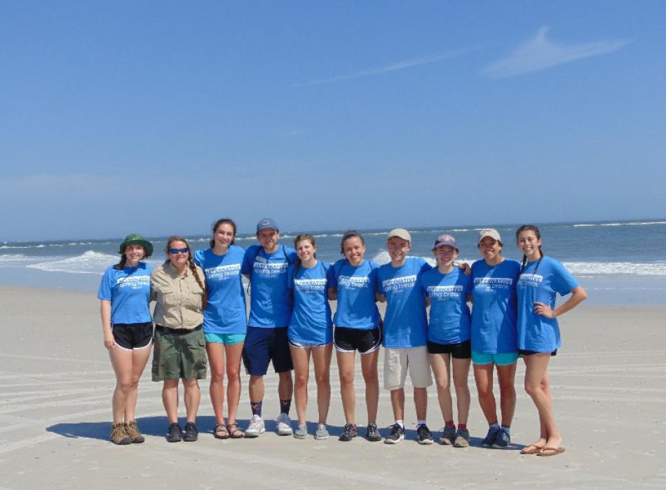 a group of college students in blue shirts and a park ranger stand arm in arm next to each other on the beach